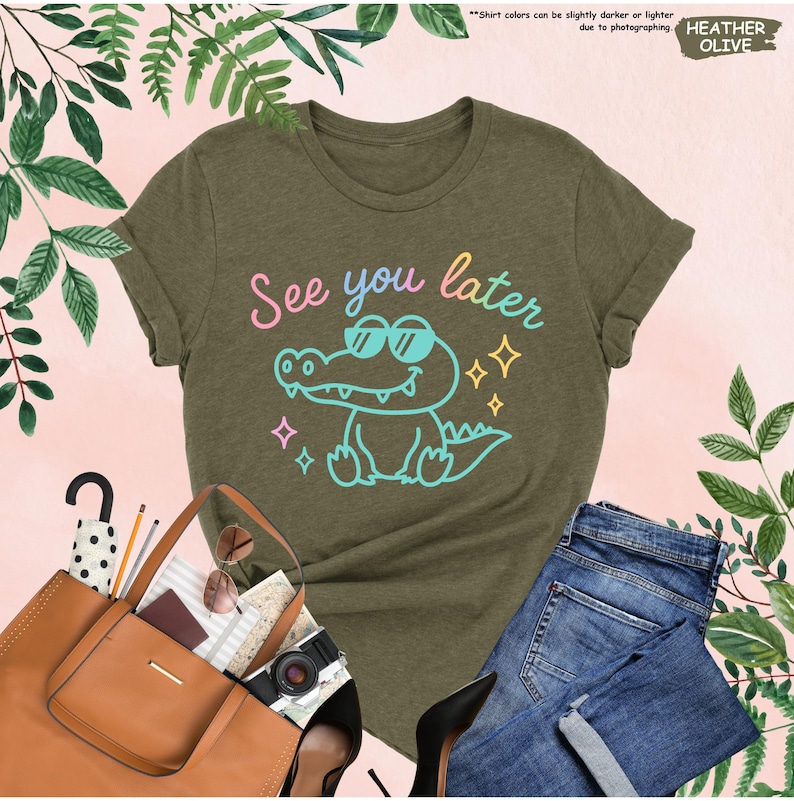 May include: Heather olive green t-shirt with the phrase "See you later" and a cartoon alligator wearing sunglasses. The shirt is paired with blue jeans, a brown tote bag, and black heels.