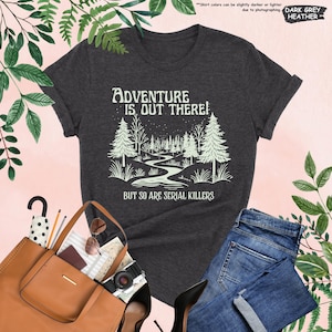 May include: Dark grey heather t-shirt with the text "ADVENTURE IS OUT THERE! BUT SO ARE SERIAL KILLERS" in white. The graphic features a forest scene with a winding river. The shirt is paired with blue jeans and a brown tote bag.