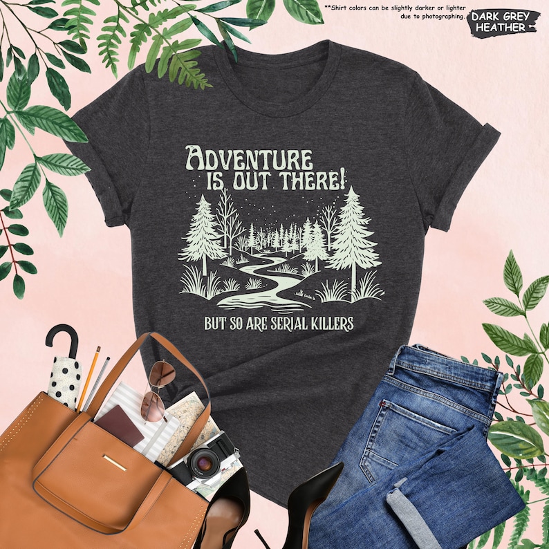 May include: Dark grey heather t-shirt with the text "ADVENTURE IS OUT THERE! BUT SO ARE SERIAL KILLERS" in white. The graphic features a forest scene with a winding river. The shirt is paired with blue jeans and a brown tote bag.