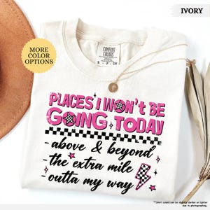 May include: Ivory-colored t-shirt with the text "PLACES I WON'T BE GOING TODAY" in pink. The shirt features a checkered flag and lightning bolt graphic. A gold necklace is draped across the shirt.