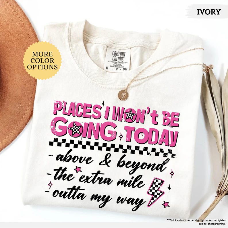 May include: Ivory-colored t-shirt with the text "PLACES I WON'T BE GOING TODAY" in pink. The shirt features a checkered flag and lightning bolt graphic. A gold necklace is draped across the shirt.