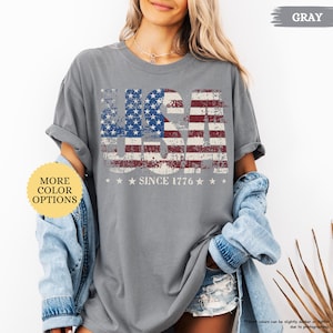 May include: Gray t-shirt with a distressed USA flag design. The design features the American flag with the text "USA" and "SINCE 1776" in white. The shirt has rolled-up sleeves and is part of a casual outfit.