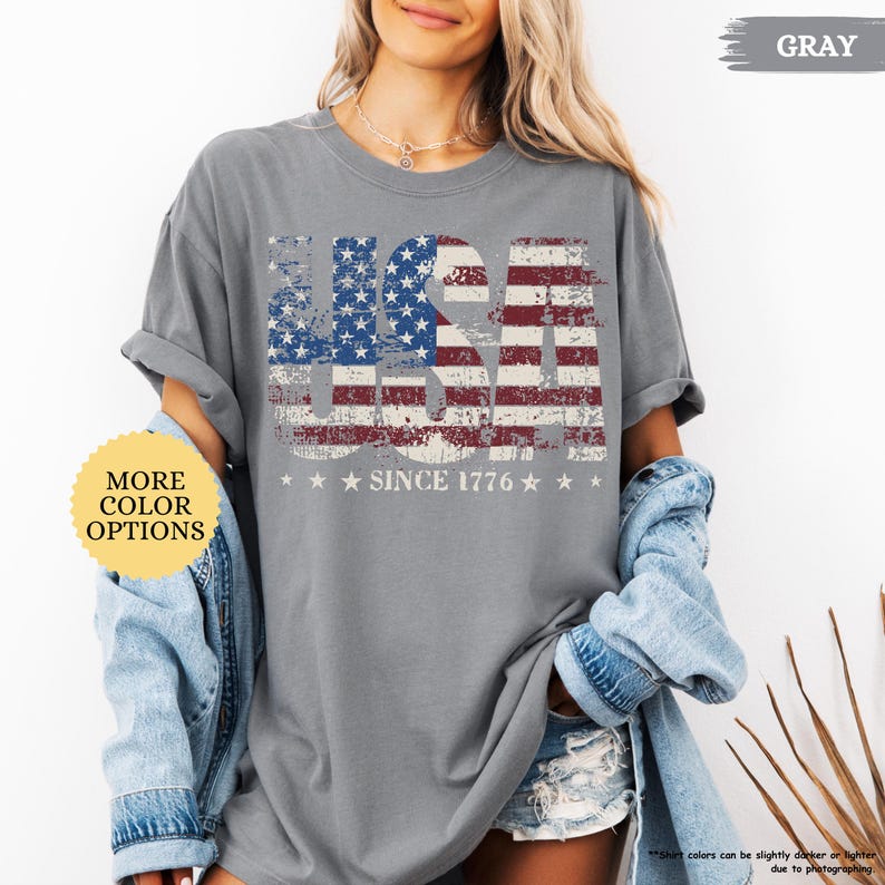May include: Gray t-shirt with a distressed USA flag design. The design features the American flag with the text "USA" and "SINCE 1776" in white. The shirt has rolled-up sleeves and is part of a casual outfit.