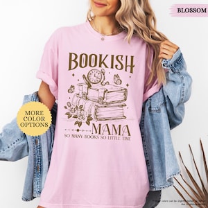 May include: A pale pink t-shirt featuring the text "BOOKISH MAMA" in brown, with a graphic of books, a clock, and butterflies. The words "SO MANY BOOKS SO LITTLE TIME" are below the graphic. The shirt has a round neck and short sleeves.