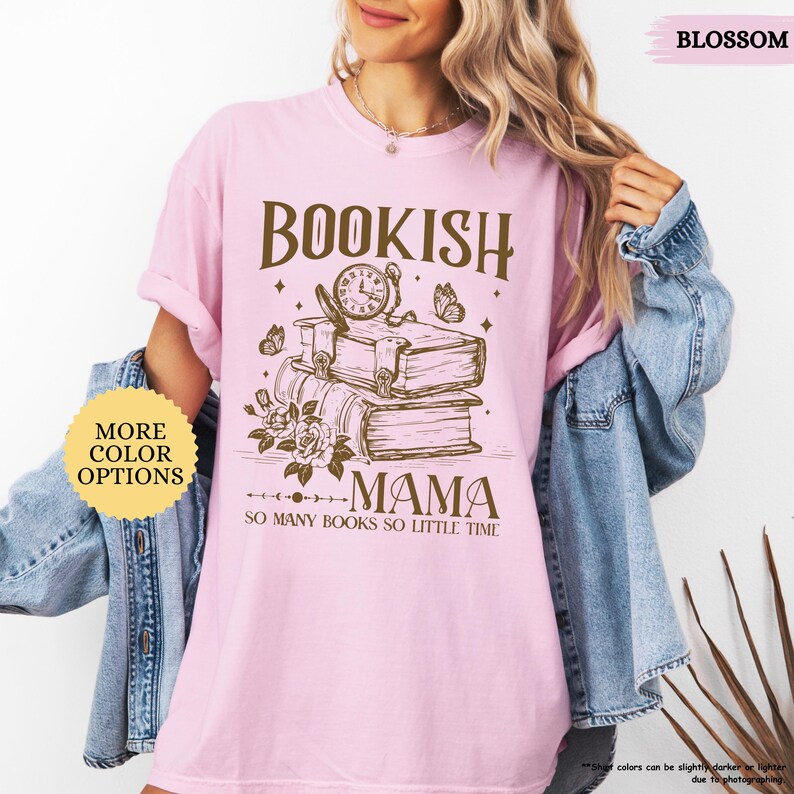May include: A pale pink t-shirt featuring the text "BOOKISH MAMA" in brown, with a graphic of books, a clock, and butterflies. The words "SO MANY BOOKS SO LITTLE TIME" are below the graphic. The shirt has a round neck and short sleeves.