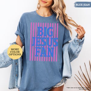 May include: Blue jean-colored t-shirt with the text "BIG JESUS FAN" in blue, set against pink vertical stripes. The shirt has rolled-up sleeves and is part of a casual wear collection. Additional text on the image includes "MORE COLOR OPTIONS" and "BLUE JEAN".