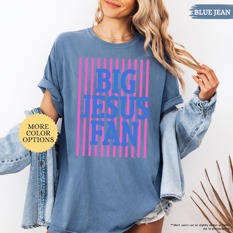 May include: Blue jean-colored t-shirt with the text "BIG JESUS FAN" in blue, set against pink vertical stripes. The shirt has rolled-up sleeves and is part of a casual wear collection. Additional text on the image includes "MORE COLOR OPTIONS" and "BLUE JEAN".
