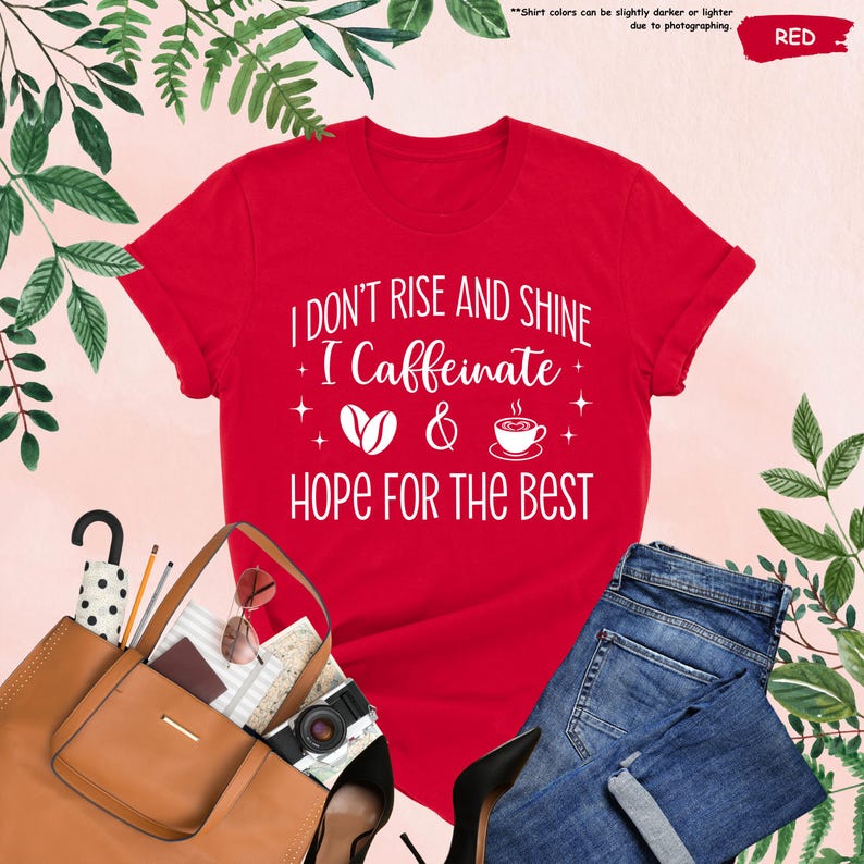May include: A red t-shirt with the text "I DON'T RISE AND SHINE I Caffeinate & HOPE FOR THE BEST" in white. The design includes coffee-related graphics. The shirt is paired with blue jeans and a brown tote bag.