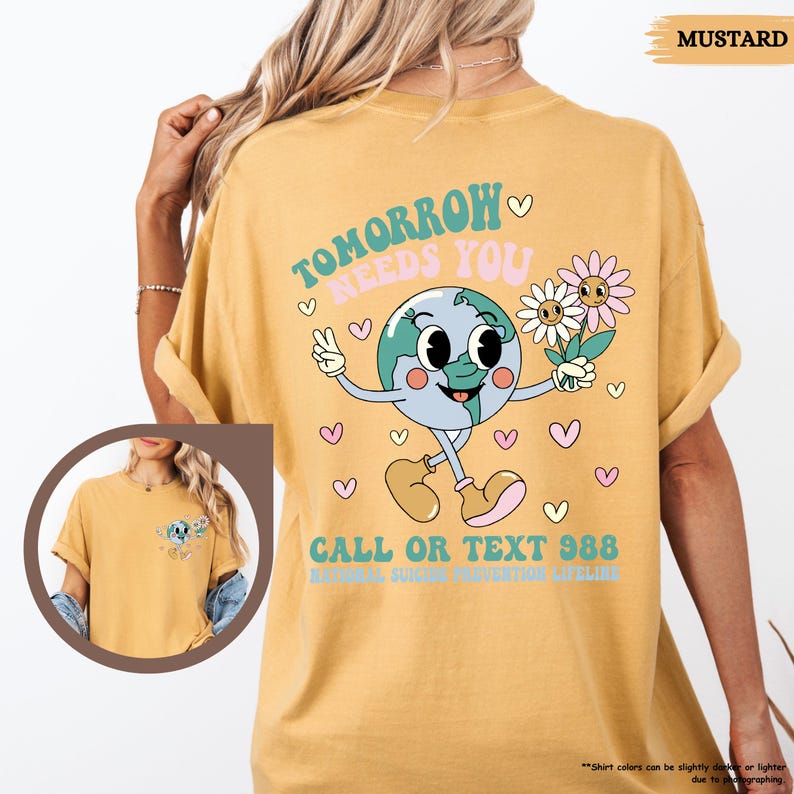 May include: Mustard-coloured t-shirt featuring a retro graphic of a smiling Earth character holding flowers, with the text "TOMORROW NEEDS YOU" and "CALL OR TEXT 988". The design includes hearts and a peace sign.