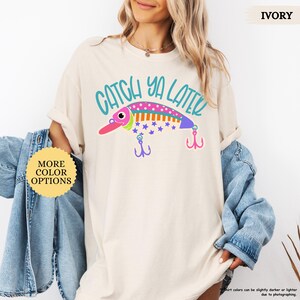 以下が含まれることがあります： アイボリーのTシャツで、カラフルな釣り用ルアーのグラフィックと「CATCH YA LATER」というティール色の文字がプリントされています。ルアーはピンク、オレンジ、紫で、水玉模様と星柄があり、紫とピンクの釣り針が2つ付いています。