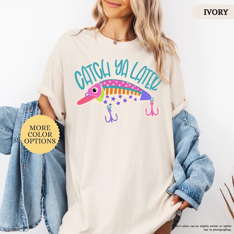 以下が含まれることがあります： アイボリーのTシャツで、カラフルな釣り用ルアーのグラフィックと「CATCH YA LATER」というティール色の文字がプリントされています。ルアーはピンク、オレンジ、紫で、水玉模様と星柄があり、紫とピンクの釣り針が2つ付いています。
