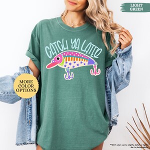 以下が含まれることがあります： ライトグリーンのTシャツで、カラフルな釣り用ルアーのグラフィックと水色のフォントで「CATCH YA LATER」という文字がプリントされています。ルアーのデザインには、ピンク、紫、オレンジの模様と星の形、そして2つのピンクの釣り針が含まれています。