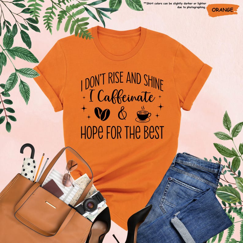 May include: Orange t-shirt with rolled sleeves featuring the text "I DON'T RISE AND SHINE I Caffeinate HOPE FOR THE BEST" in black. The design includes coffee beans and a coffee cup. The shirt is paired with blue jeans and accessories.