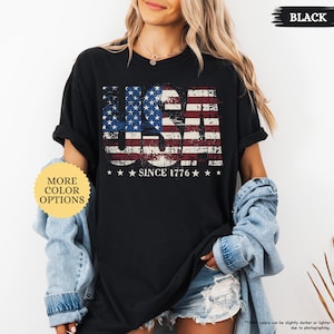 May include: Black t-shirt with a distressed USA flag design. The word "USA" is prominently displayed, with the flag's stars and stripes incorporated into the letters. Below the design, the text reads "SINCE 1776".