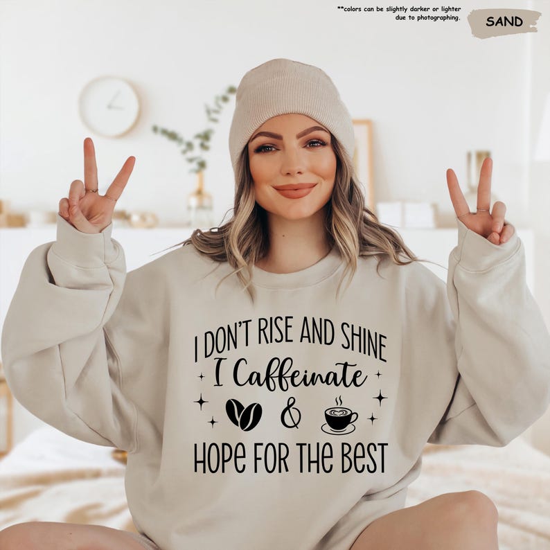 May include: A sand-colored sweatshirt with the text "I DON'T RISE AND SHINE I Caffeinate & HOPE FOR THE BEST" in black. The design includes coffee beans, a coffee cup, and stars. The model is wearing a sand-colored beanie.