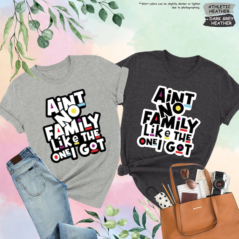 May include: Two t-shirts, one athletic heather and one dark grey heather, with the text "Ain't No Family Like The One I Got" in a colorful, bold font. The athletic heather shirt is paired with blue jeans. A brown tote bag is next to the dark grey shirt.
