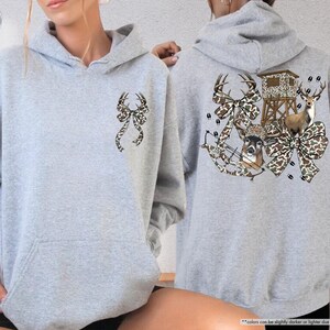 May include: Sport grey hoodie with a front pocket and a hood. The front features a bow with deer antlers in a camouflage pattern. The back displays a hunting scene with deer, a bow, and a deer stand, all in a camouflage bow design.