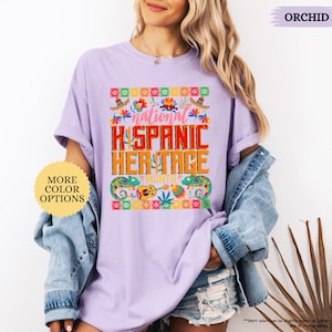 Peut inclure: T-shirt lilas avec l'inscription "national HISPANIC HERITAGE MONTH" dans un design color&eacute;. Le motif comprend des sombreros, des fleurs et des cam&eacute;l&eacute;ons. Le t-shirt a &eacute;galement un bouton "MORE COLOR OPTIONS".