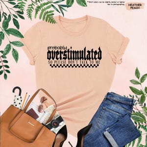 May include: A peach coloured t-shirt with black text that reads "probably overstimulated so please don't talk to me" with a black and white chequered pattern underneath the text.
