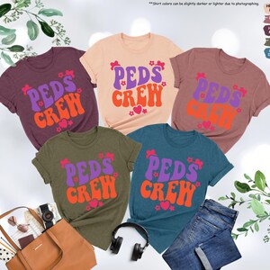 May include: Five t-shirts in different colors with the text "PEDS CREW" in a retro font. The text is in purple and orange with pink hearts and flowers.
