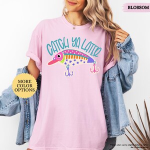 以下が含まれることがあります： 淡いピンク色の半袖Tシャツ。カラフルな釣り用ルアーのグラフィックと、ティール色の文字で「CATCH YA LATER」とプリントされています。ルアーのデザインには、ピンク、オレンジ、青の色合いで水玉模様、ストライプ、星柄が描かれています。