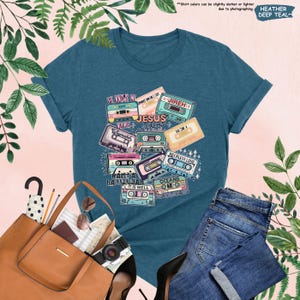 May include: A teal t-shirt featuring a graphic of vintage cassette tapes with religious phrases like "Jesus" and "Waymaker." The shirt is paired with blue jeans, a brown tote bag, and black heels. The text "HEATHER DEEP TEAL" is in the upper right corner.