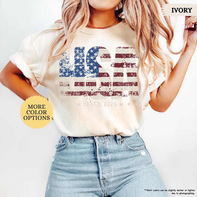 May include: Ivory-colored t-shirt featuring a distressed USA flag design in blue, white, and red. The text "USA" is prominently displayed, with "SINCE 1776" printed below. The shirt is paired with blue jeans.