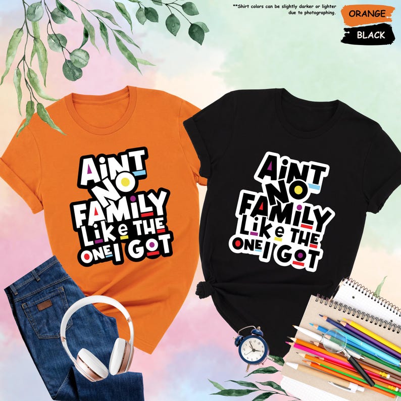 May include: Two t-shirts, one orange and one black, with the text "Aint No Family Like The One I Got" in a colorful, bold font. The orange shirt is paired with blue jeans and white headphones. The black shirt is styled with a small alarm clock and colored pencils.