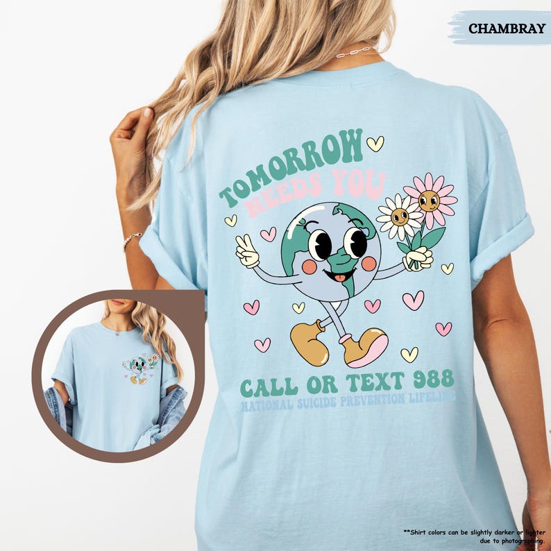 May include: Pale blue t-shirt featuring the text "TOMORROW NEEDS YOU" and a cartoon earth character holding flowers. The shirt also has the text "CALL OR TEXT 988" and "NATIONAL SUICIDE PREVENTION LIFELINE".