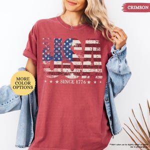 May include: Crimson-colored t-shirt with a distressed USA flag design. The design includes the text "USA" in a patriotic font, with the stars and stripes. Below the flag design is the text "SINCE 1776".