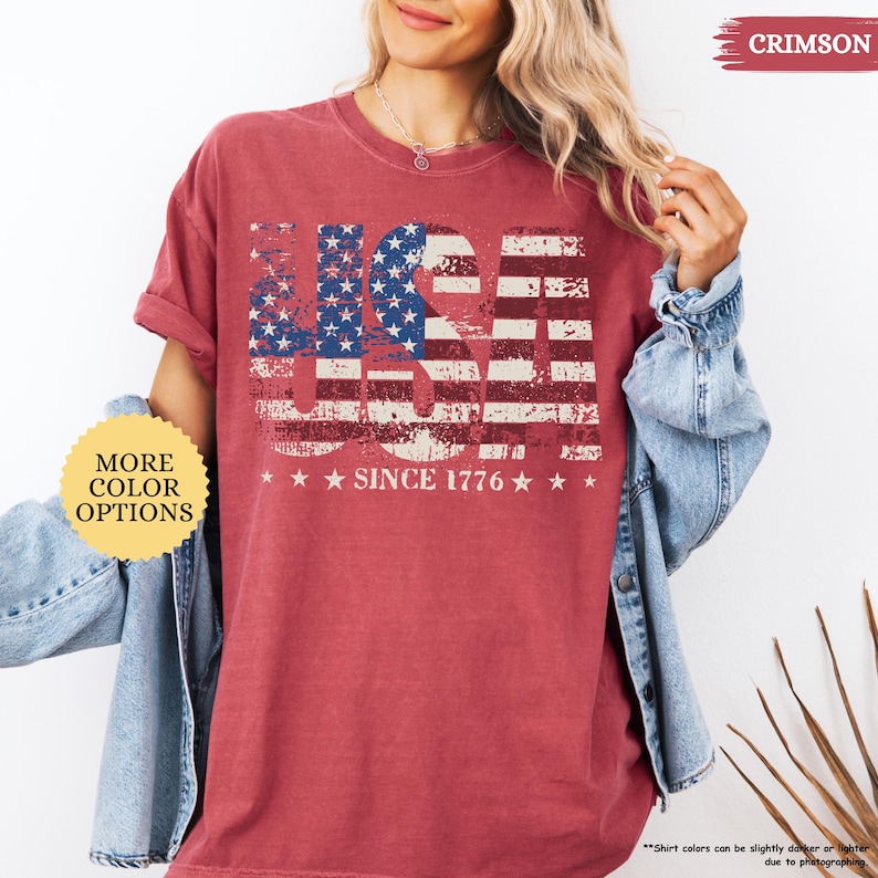 May include: Crimson-colored t-shirt with a distressed USA flag design. The design includes the text "USA" in a patriotic font, with the stars and stripes. Below the flag design is the text "SINCE 1776".