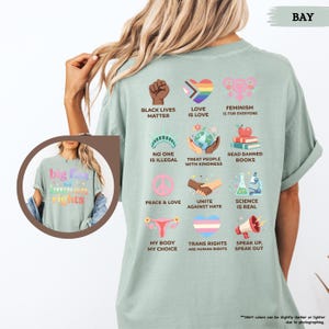 May include: A light green t-shirt with various graphic designs and text promoting social justice and equality. The shirt includes phrases like "Black Lives Matter," "Love is Love," and "Trans Rights are Human Rights."