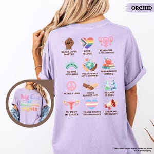 May include: Orchid-colored t-shirt with various social justice graphics and text. The shirt features phrases like "Black Lives Matter," "Love is Love," and "Feminism is for Everyone." Additional graphics include a rainbow heart, a globe, and a peace sign.