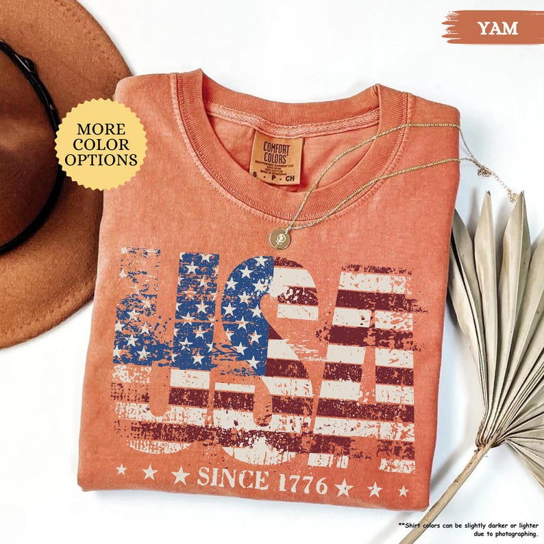May include: A coral-colored USA graphic t-shirt with a distressed American flag design. The shirt features the text "USA" in bold letters, with the flag's stars and stripes incorporated into the design. The text "SINCE 1776" is printed below the graphic.