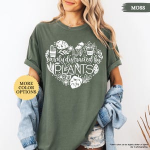 Pu&ograve; includere: Una maglietta verde con un design a forma di cuore bianco con il testo "easily distracted by PLANTS" e una variet&agrave; di illustrazioni di piante. La maglietta ha una vestibilit&agrave; rilassata e un collo rotondo.