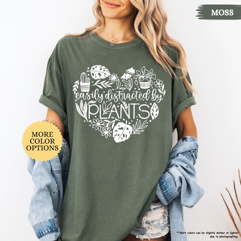 Pu&ograve; includere: Una maglietta verde con un design a forma di cuore bianco con il testo "easily distracted by PLANTS" e una variet&agrave; di illustrazioni di piante. La maglietta ha una vestibilit&agrave; rilassata e un collo rotondo.