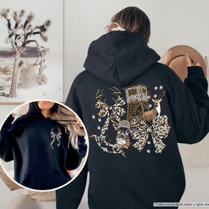 May include: Black hooded sweatshirt with a hunting-themed graphic on the back. The design features a deer, a hunting blind, and camouflage bows. The front of the sweatshirt has a small bow design. The sweatshirt is made of a soft material.