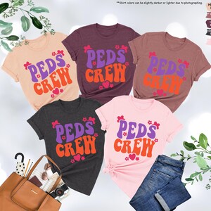 May include: Five t-shirts in different colors with the text "PEDS CREW" in a retro font. The text is in purple, orange, and pink. The shirts are a heather peach, heather mauve, heather maroon, dark grey heather, and pink.