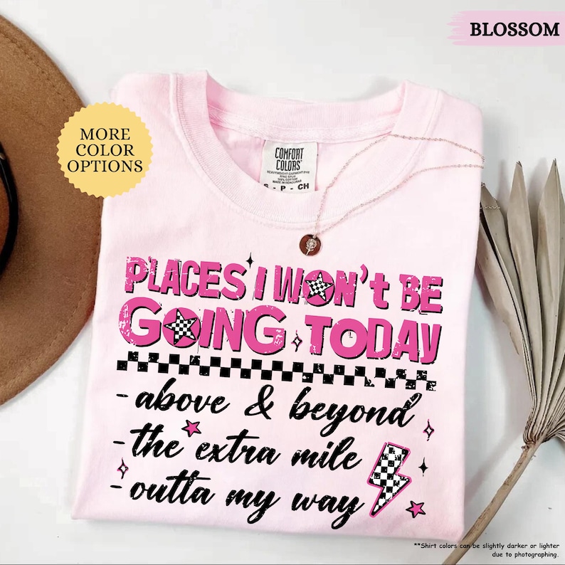 May include: A light pink t-shirt with the text "PLACES I WON'T BE GOING TODAY" in a distressed pink font. The shirt also features the phrases "above & beyond", "the extra mile", and "outta my way". A gold necklace is draped over the shirt.