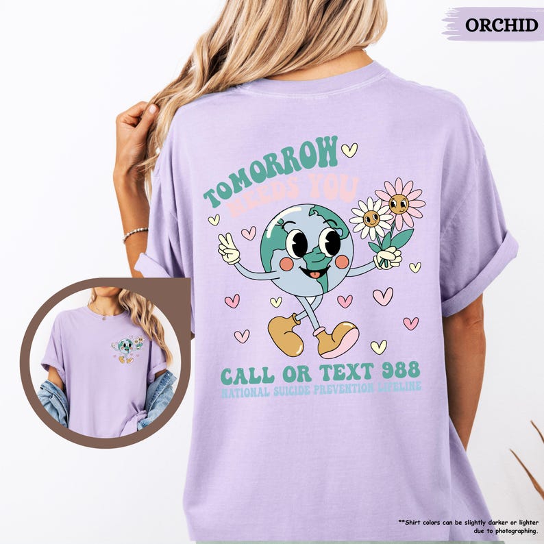 May include: Lilac t-shirt featuring a cartoon globe character with flowers and a peace sign. Text on the shirt reads "TOMORROW LOVES YOU" and "CALL OR TEXT 988 NATIONAL SUICIDE PREVENTION LIFELINE".