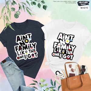 May include: Two t-shirts, one navy and one white, with the text "Ain't No Family Like The One I Got" in a colorful, bold font. The navy shirt is paired with blue jeans. The white shirt is next to a brown tote bag.