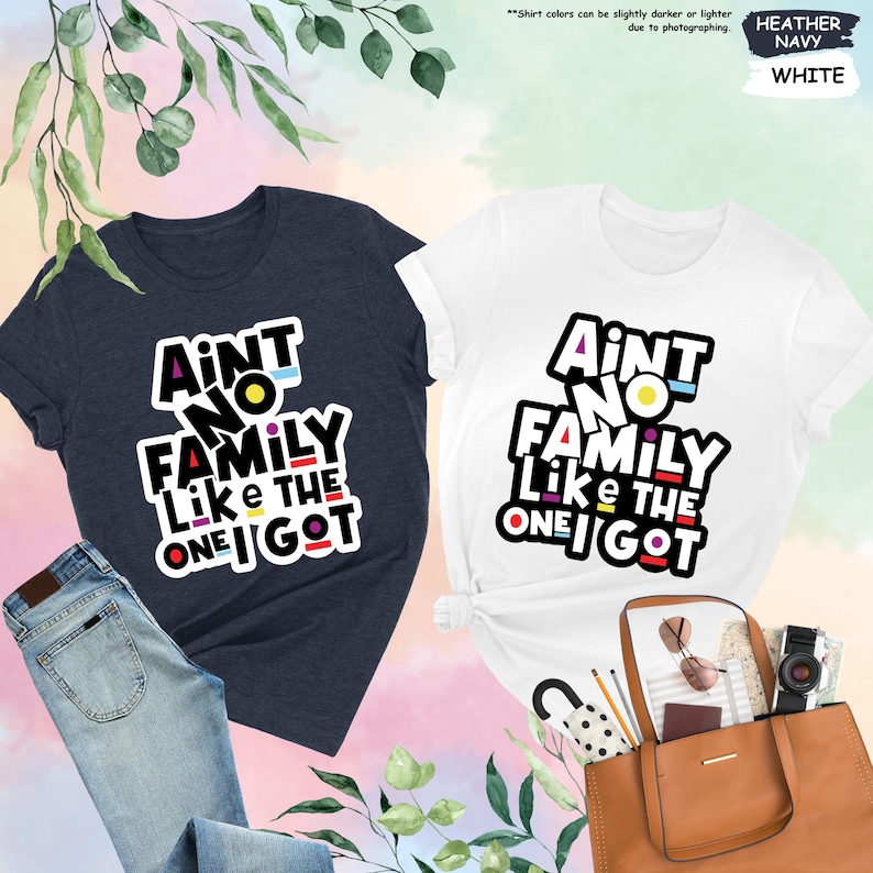 May include: Two t-shirts, one navy and one white, with the text "Ain't No Family Like The One I Got" in a colorful, bold font. The navy shirt is paired with blue jeans. The white shirt is next to a brown tote bag.
