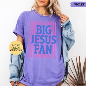 May include: A violet t-shirt with the text "BIG JESUS FAN" in blue, framed by pink vertical stripes. The shirt has rolled-up sleeves and a relaxed fit. A small sticker on the left says "MORE COLOR OPTIONS".