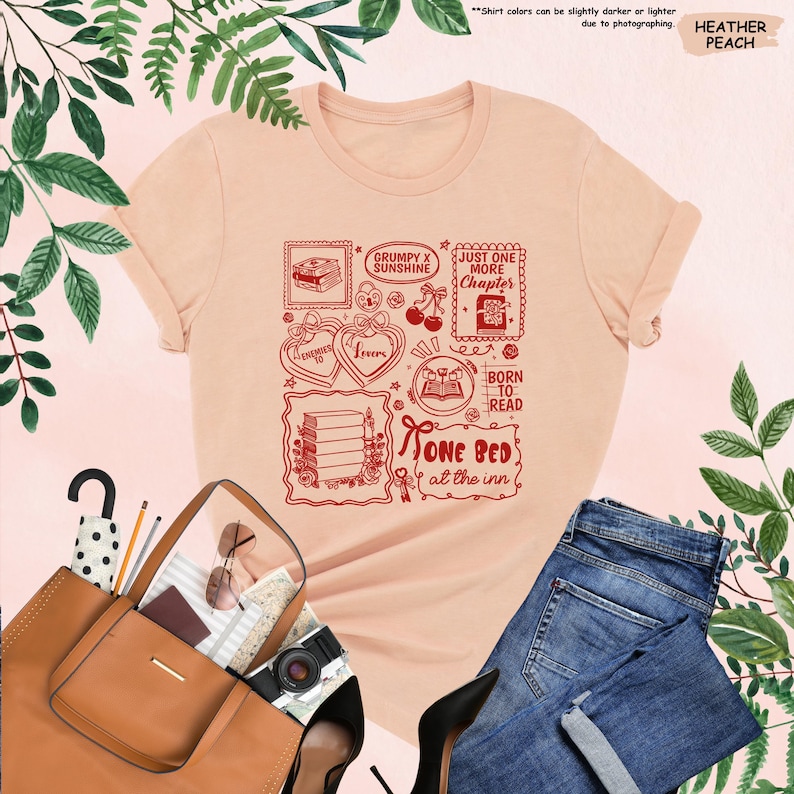 May include: Heather peach t-shirt with a red graphic design featuring book-themed illustrations and text. The design includes phrases like "Grumpy x Sunshine," "Just One More Chapter," and "Born to Read." The shirt is paired with blue jeans and a brown handbag.