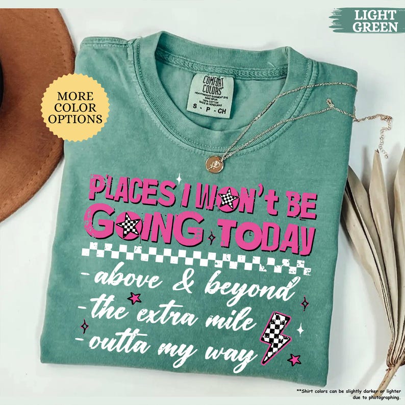 May include: A light green t-shirt with the text "PLACES I WON'T BE GOING TODAY" in pink. The shirt has a crew neck and short sleeves. A silver necklace with a pendant is on the shirt.