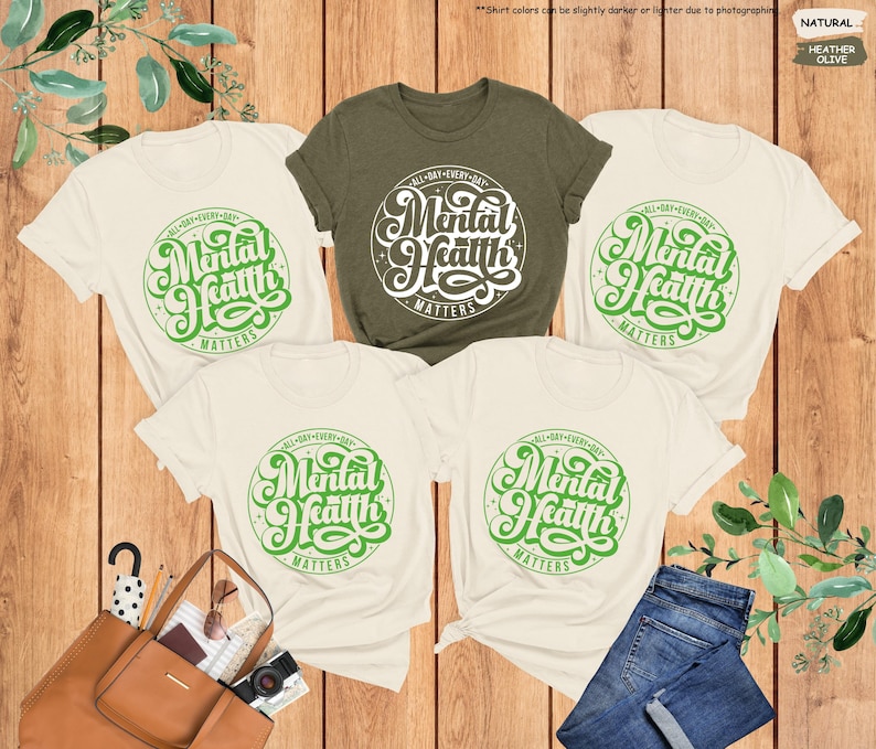 May include: Four t-shirts with the text 'All Day Every Day Mental Health Matters' in green lettering on a white circle. The shirts are laid out on a wooden surface with green plants and a brown leather bag in the background. The shirts are available in natural and heather olive colors.