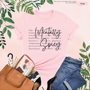 K&ouml;nnte beinhalten: Ein rosa T-Shirt mit den Worten "Mentally Spicy" in einer stilvollen Schriftart. Das Shirt listet auch mehrere Adjektive wie "Edgy", "Sassy" und "Fearless" auf. Das Shirt wird mit einer blauen Jeans, einer braunen Handtasche und schwarzen Abs&auml;tzen kombiniert.