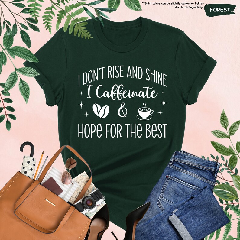 May include: A dark green t-shirt with the text "I DON'T RISE AND SHINE I Caffeinate HOPE FOR THE BEST" in white. The shirt is paired with blue jeans, a brown purse, and black heels. The background has green foliage.
