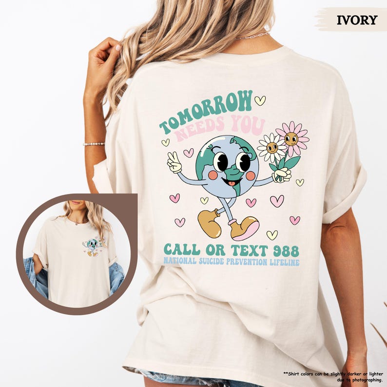May include: Cream-coloured t-shirt with a cartoon globe design and the text "Tomorrow Needs You" and "Call or Text 988". The globe character holds flowers and a peace sign. A casual, unisex style.
