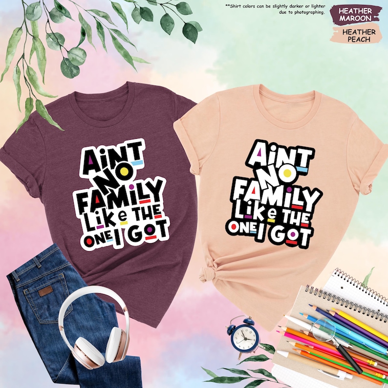 May include: Two t-shirts, one heather maroon and one heather peach, with the text "Ain't No Family Like The One I Got" in a bold, colorful design. The shirts are displayed with a pair of blue jeans, white headphones, a small clock, and colored pencils.